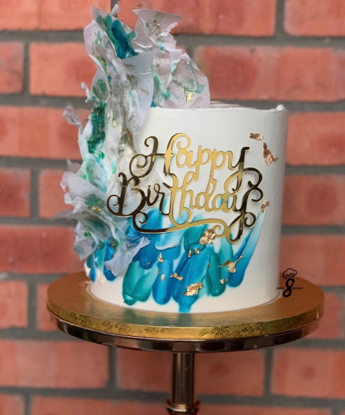 Bolo branco com pinceladas de azul e turquesa, folha de ouro e topper “Happy Birthday” sobre pedestal dourado